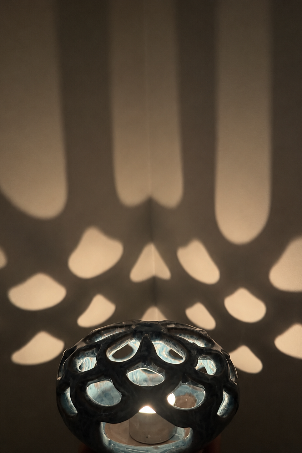 A close-up of a dark, textured metal decorative lamp shade with cutout patterns, casting a geometric shadow on a wall behind it.